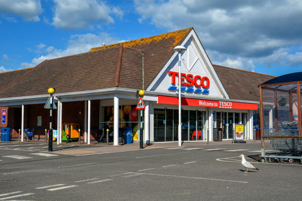 The entrance of a Tesco supermarket in Honiton, Devon, under a bright blue sky with white clouds